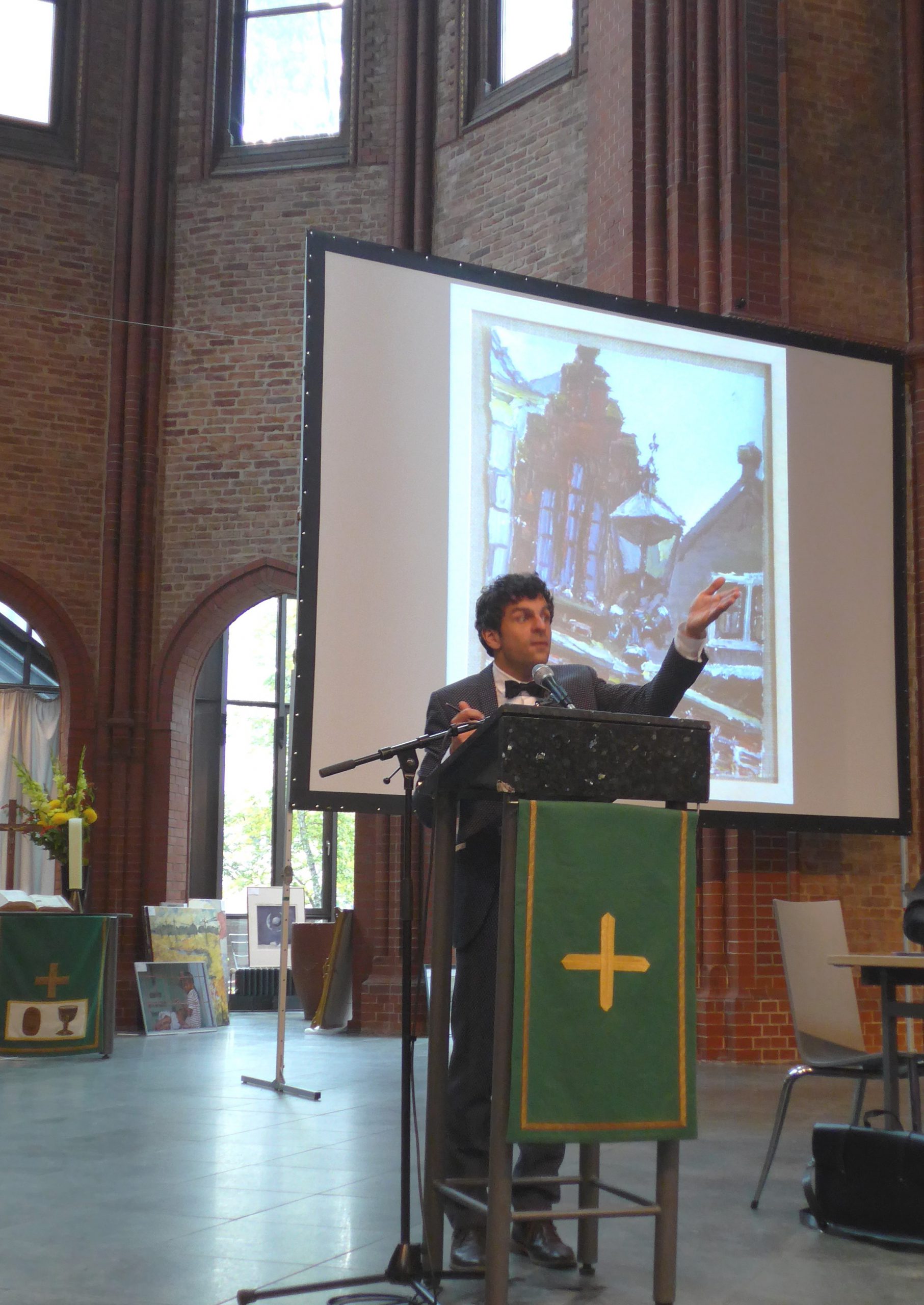 Ein Mann im Anzug spricht an einem Podium in einer Kirche mit Backsteinmauern, einem großen Kreuz und hohen Fenstern. Hinter ihm zeigt ein Bildschirm das Bild eines Gemäldes oder einer Fotografie von Gebäuden.