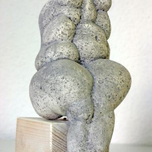 Eine Steinskulptur mit einem stilisierten, abstrakten weiblichen Torso mit übertriebenen Kurven, montiert auf einem hellen Holzsockel und vor einem hellen Hintergrund.