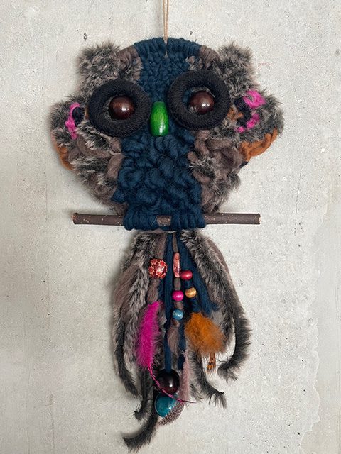 Ein handgefertigter Eulen-Wandbehang aus Garn, Stoff und Perlen mit großen Knopfaugen, einem grünen Schnabel, einer Stange und darunter hängenden federartigen Verzierungen. Der Hintergrund ist eine schlichte, helle Wand.