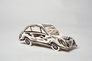 Eine Keramikskulptur eines Autos mit schwarzen und braunen handgezeichneten Mustern und Linien auf weißem Hintergrund, die ihm ein stilisiertes, künstlerisches Aussehen verleihen. Der Hintergrund ist einfarbig und hell gefärbt.