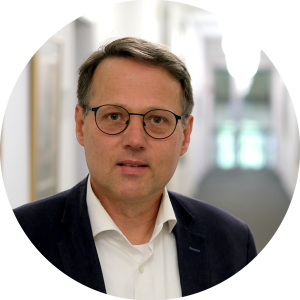 Ein Mann mittleren Alters mit kurzen braunen Haaren, Brille und einem dunklen Blazer über einem weißen Hemd steht in einem Flur mit unscharfem Hintergrund.