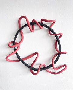 Ein schwarzer, kreisförmiger Ring mit einer dicken, gewellten roten Linie, die sich entlang des Randes hin und her schlängelt, montiert auf einer schlichten weißen Wand.