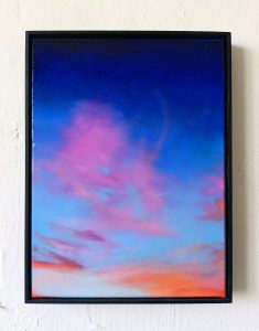 Ein gerahmtes Gemälde eines farbenfrohen Himmels, der oben tiefblau ist und unten in rosa und orangefarbene Wolken übergeht, vor einer schlichten weißen Wand.