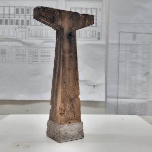 Eine Betonskulptur in Form eines umgekehrten "T" steht auf einer weißen Fläche, im Hintergrund sind Architekturzeichnungen zu sehen. Die Skulptur hat eine raue Textur und einen Sockel aus Metallgewebe.