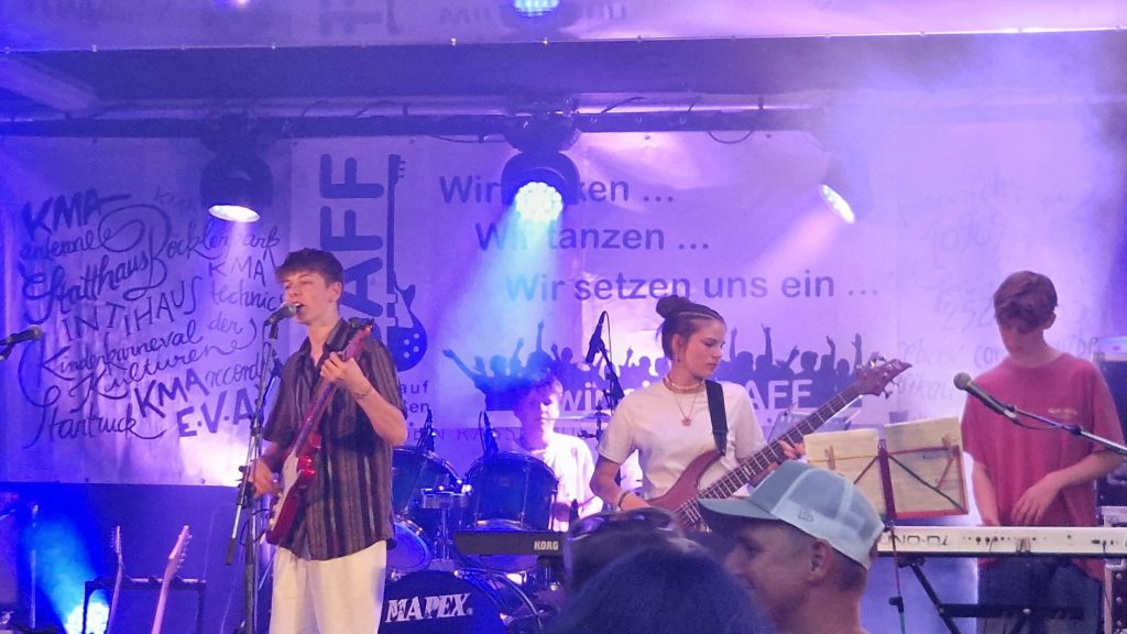 Eine Band tritt auf der Bühne mit Gitarren, Schlagzeug und Keyboard auf. Vier Musiker spielen unter lila Lichtern, während die Menge zusieht. Auf einem Transparent hinter ihnen steht "Wir setzen uns ein..." auf Deutsch.