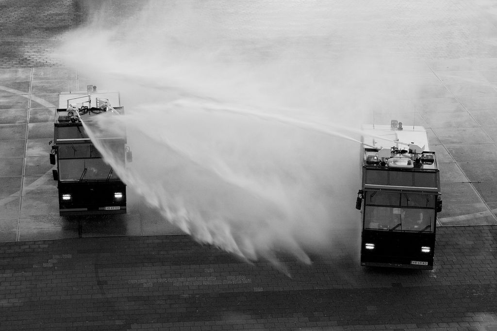 Zwei große Fahrzeuge versprühen aus den auf ihren Dächern montierten Schläuchen kräftige Wasserstrahlen, die dichten Nebel erzeugen. Die Szene scheint auf einer gepflasterten Fläche zu spielen und ist in Schwarzweiß aufgenommen.