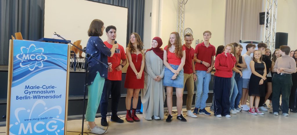 Eine Gruppe von Schülern steht in einer Reihe auf der Bühne, viele tragen rote oder beigefarbene Kleidung. Eine Frau hält ein Mikrofon und spricht mit einem Jungen. Auf der linken Seite ist ein Podium mit dem Logo des Marie-Curie-Gymnasiums Berlin-Wilmersdorf zu sehen.