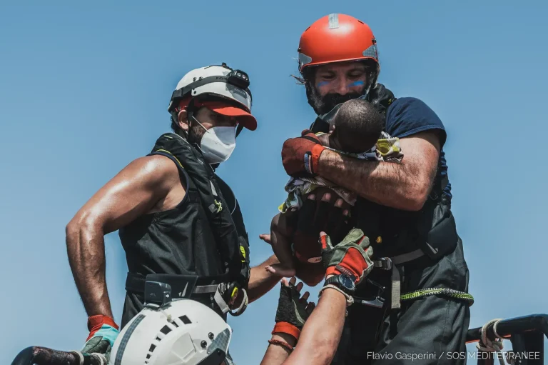 Zwei Rettungskräfte mit Helmen und Sicherheitsausrüstung halten und helfen einem kleinen Kind, das von einer anderen Person hochgehoben wird, vor einem klaren blauen Himmel. Die Szene vermittelt den Eindruck einer Notsituation.