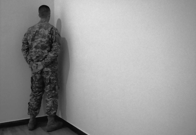 Eine Person in Militäruniform steht in einer Ecke und hat die Hände hinter dem Rücken verschränkt. Der Raum hat schlichte Wände und einen Holzboden, was eine strenge und minimalistische Atmosphäre schafft.