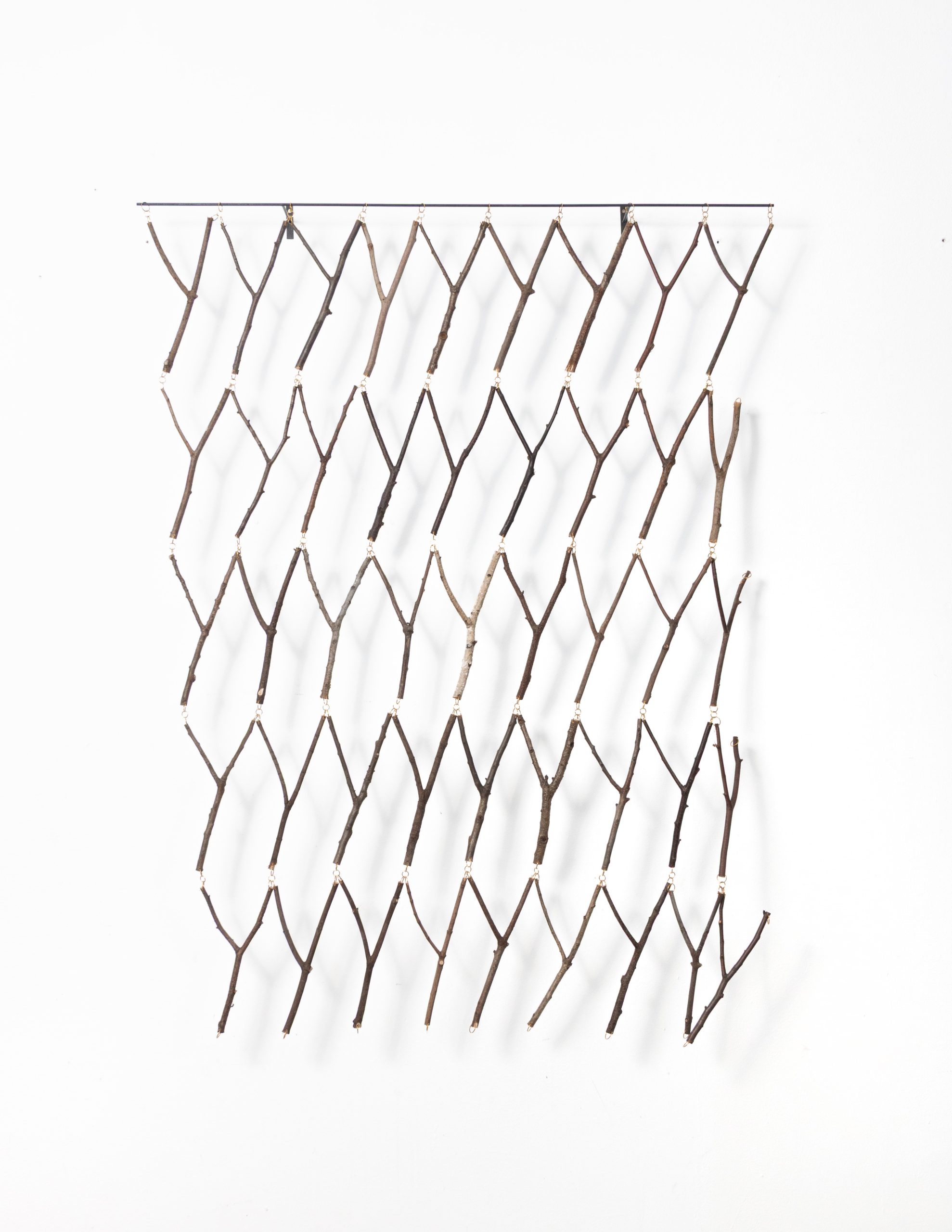 Ein minimalistischer Wandbehang aus mehreren Reihen kleiner, Y-förmiger Zweige, die mit Schnur zusammengebunden sind und an einer horizontalen Stange vor einem weißen Hintergrund hängen.
