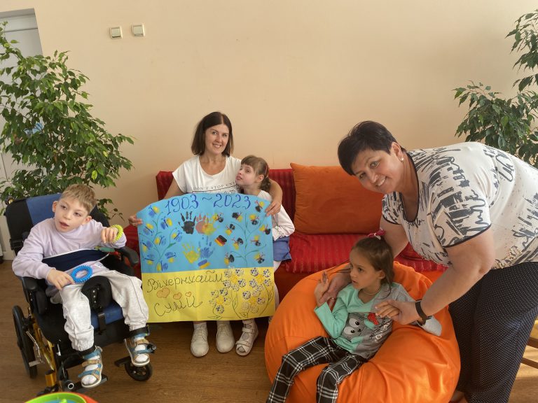 Vier Kinder und zwei Frauen posieren drinnen mit einem bunten, handgefertigten Plakat mit der Aufschrift 1903-2023. Ein Kind sitzt in einem Rollstuhl, ein anderes auf einem orangefarbenen Sitzsack. Die Gruppe sieht fröhlich und engagiert aus.