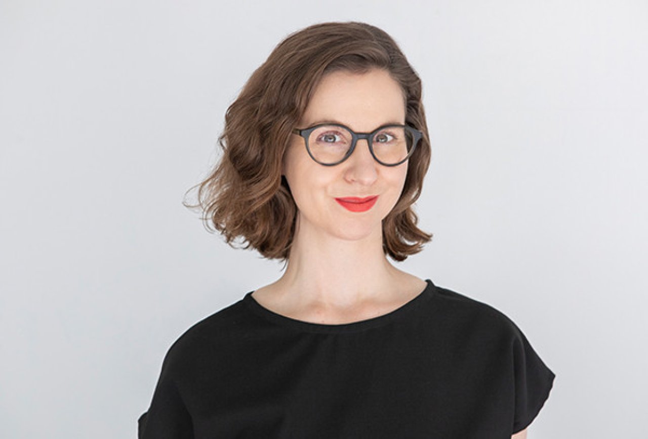 Eine Frau mit kurzen, gewellten braunen Haaren, die eine Brille, roten Lippenstift und ein schwarzes Oberteil trägt, lächelt in die Kamera vor einem schlichten weißen Hintergrund.