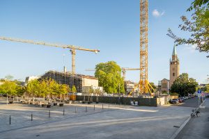 Neben einer von Bäumen gesäumten Straße ist eine Baustelle mit Kränen und Gerüsten zu sehen. Im Hintergrund ist ein hoher Kirchturm mit grüner Spitze unter einem klaren blauen Himmel zu sehen.