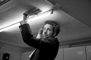 Ein Mann im Anzug steht auf einer Fläche und schaut in die Kamera, während er eine Deckenleuchte verstellt. Eine seiner Hände ist behandschuht. Das Foto ist in schwarz-weiß gehalten.