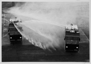 Zwei große Fahrzeuge versprühen aus montierten Schläuchen kräftige Wasserstrahlen, die eine Nebelwolke erzeugen. Die Szene scheint auf einem nassen Bürgersteig zu spielen, möglicherweise während einer Übung zur Kontrolle von Menschenmengen oder einer Übung.