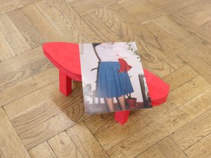 Ein kleiner, roter, abstrakter Holztisch steht auf einem Parkettboden. Darauf befindet sich ein Foto, das eine Person in weißem Hemd und blauem Rock zeigt, von der nur der Unterkörper zu sehen ist. Im Hintergrund stehen Kübelpflanzen.