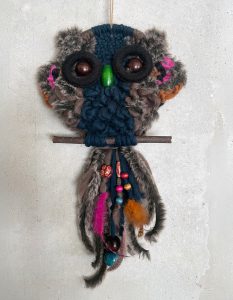 Ein handgefertigter Eulen-Wandbehang aus Garn, Kunstfell und Perlen, mit Knopfaugen und grünem Schnabel, auf einem Stock sitzend, verziert mit bunten Federn und Perlen, vor einer hellen, strukturierten Wand.