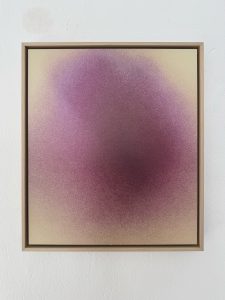 Ein gerahmtes abstraktes Kunstwerk mit einem weichen Farbverlauf aus lila und rosa Tönen, die in einen hellbeigen Hintergrund übergehen und einen nebelartigen Effekt erzeugen. Der Rahmen ist einfach und holzfarben vor einer weißen Wand.