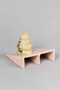 Eine kleine, gelbliche, gewundene Keramikskulptur steht auf einer dreieckigen, dreibeinigen, rosafarbenen Plattform vor einem schlichten grauen Hintergrund.