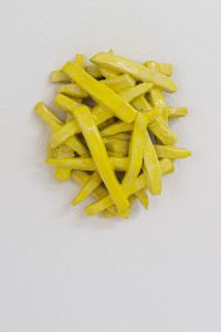 Eine Keramikskulptur, die an einen Haufen gelber Pommes frites erinnert, ist an einer weißen, strukturierten Wand angebracht.