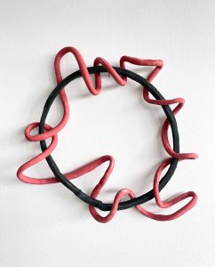 Ein kreisförmiger schwarzer Ring hängt an einer weißen Wand, und ein wellenförmiges rotes Element schlängelt sich unregelmäßig um und durch den Ring. Die Skulptur wirft subtile Schatten auf die Wand hinter ihr.