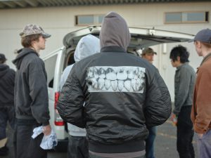 Eine Gruppe von Personen steht neben einem offenen Kofferraum. Die Person in der Mitte trägt eine schwarze Jacke mit einer großen Grafik von Zähnen auf dem Rücken. Die meisten tragen Freizeitkleidung und Hüte. Die Szene scheint im Freien zu spielen.