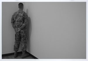 Eine Person in Militäruniform steht mit hinter dem Rücken verschränkten Händen in der Ecke eines weitgehend leeren Raums.