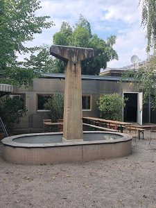 Ein hoher, T-förmiger Betonbrunnen steht in einem leeren, runden Becken in einem sandigen Innenhof, umgeben von Bäumen, Bänken und einem Gebäude mit großen Fenstern und einer offenen Tür.