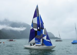 Ein Segelboot mit einem geometrischen blauen, schwarzen und grauen Segel fährt über einen ruhigen See, mit nebligen Bergen und anderen Booten im Hintergrund unter einem bedeckten Himmel.