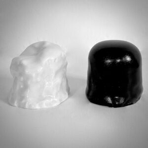 Zwei kuppelförmige Bonbons liegen nebeneinander auf einer weißen Fläche; das linke ist glänzend weiß und hat eine raue, unregelmäßige Oberfläche, das rechte ist glatt und glänzend schwarz.