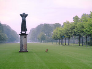 In einem grasbewachsenen Park steht eine große Frauenstatue auf einem Sockel, neben ihr sitzt ein kleiner Hund. Bäume säumen beide Seiten der Freifläche unter einem nebligen, pastellfarbenen Himmel.