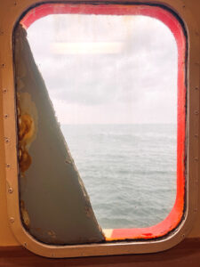 Blick auf ein bewölktes, graues Meer durch ein rechteckiges Bullauge, dessen Rahmen Rostspuren und abblätternde Farbe aufweist, gesehen vom Inneren eines Schiffes oder Bootes aus.