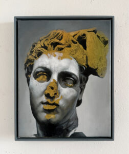 Ein gerahmtes Gemälde, das den Kopf einer klassischen Statue zeigt, wobei Teile des Gesichts und der Haare mit goldener und weißer Farbe übermalt sind; das Bild ist vor einem grauen Hintergrund dargestellt.