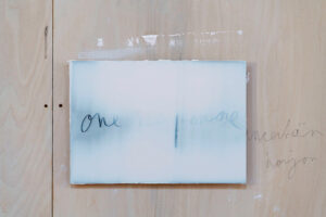 Ein rechteckiges Kunstwerk mit der Aufschrift one more in Schreibschrift, angebracht an einer Sperrholzwand, auf der rechts mit Bleistift die Worte uncertain horizon stehen.