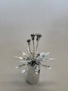 Eine glänzende Metallskulptur einer distelartigen Blume mit stacheligen Blütenblättern und Stielen, präsentiert vor einem schlichten, hellen Hintergrund.
