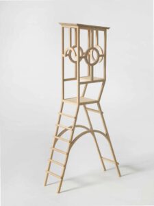 Eine kleine Holzskulptur mit einer Leiter, die zu einer geometrischen, offenen Struktur aus kreisförmigen und rechteckigen Formen hinaufführt, vor einem schlichten weißen Hintergrund.