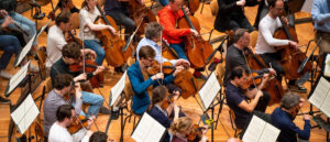 Eine Aufnahme aus der Vogelperspektive eines vielfältigen Orchesterensembles, das während einer Probe auf Celli und Violinen spielt.