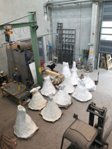 Eine Person arbeitet an einem Tisch in einer Werkstatt, in der mehrere in weiße Plastikfolie eingewickelte Skulpturen stehen, darunter mehrere glockenförmige Figuren und ein großer schwarzer Vogel.